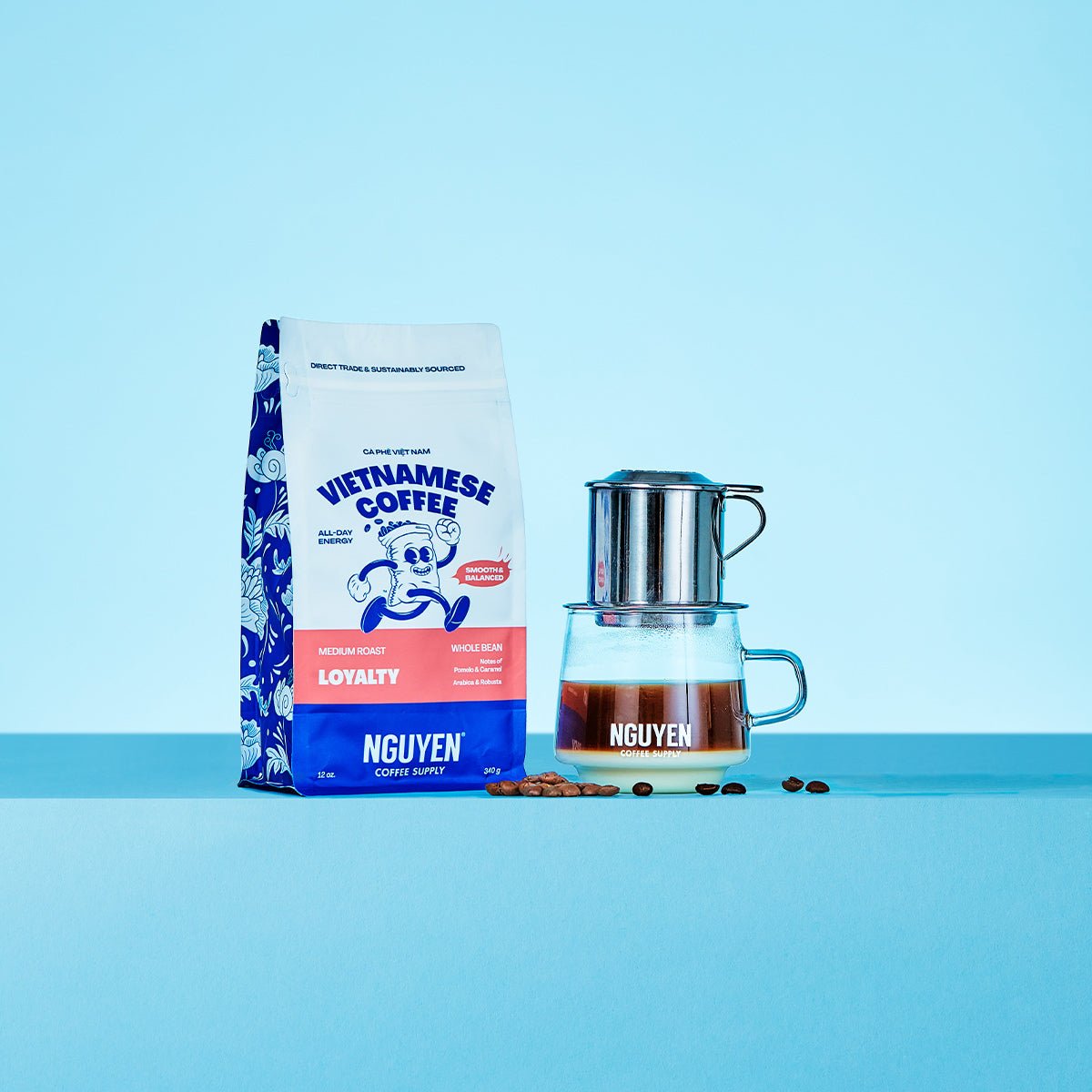 A bag of Nguyen Vietnamese Coffee Loyalty medium roast coffee beans stands next to a traditional phin filter dripping over a glass cup filled with brewed coffee and sweetened condensed milk, with a few coffee beans scattered in between, on a light blue background.
