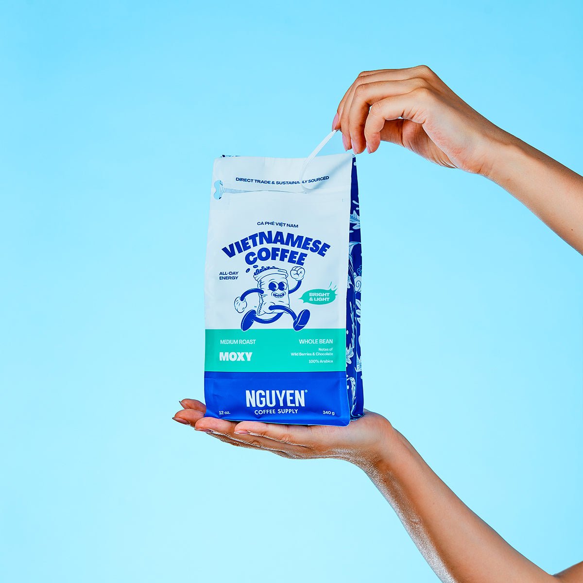 A person holds a bag of Nguyen Vietnamese Coffee Moxy against a solid light blue background. The bag is white with blue branding and a small green label, with one hand pulling the tab and the other supporting the bottom.